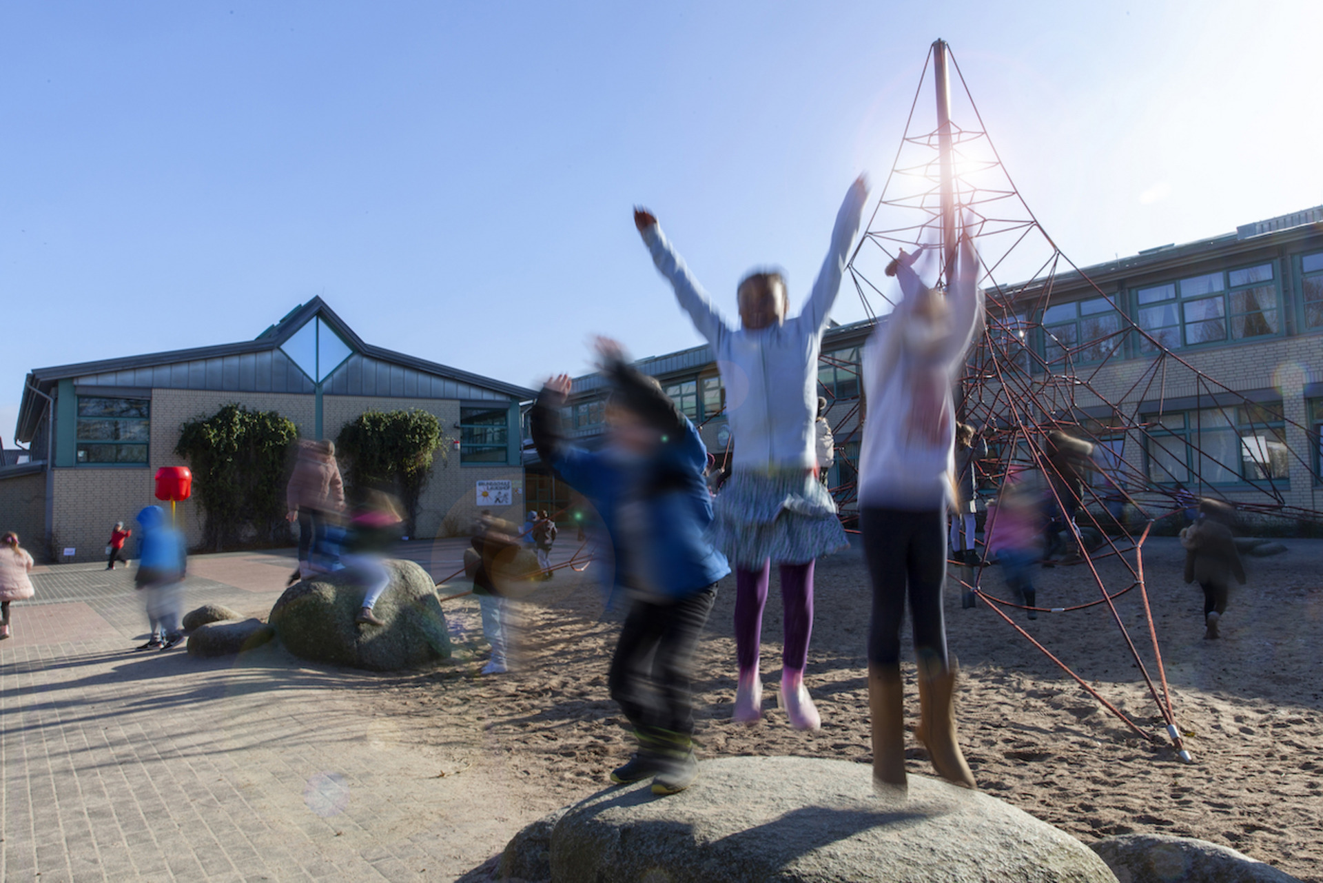 Kinder spielen ausgelassen auf einem Schulhof. Im Vordergrund springen mehrere Kinder mit erhobenen Armen von Felsen, ihre Bewegungen sind verschwommen. Im Hintergrund sieht man ein Schulgebäude mit großen Fenstern und eine rote Kletterstruktur. Die Szene strahlt Energie und Freude aus.