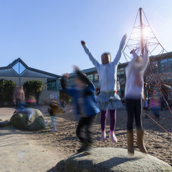 Kinder spielen ausgelassen auf einem Schulhof. Im Vordergrund springen mehrere Kinder mit erhobenen Armen von Felsen, ihre Bewegungen sind verschwommen. Im Hintergrund sieht man ein Schulgebäude mit großen Fenstern und eine rote Kletterstruktur. Die Szene strahlt Energie und Freude aus.