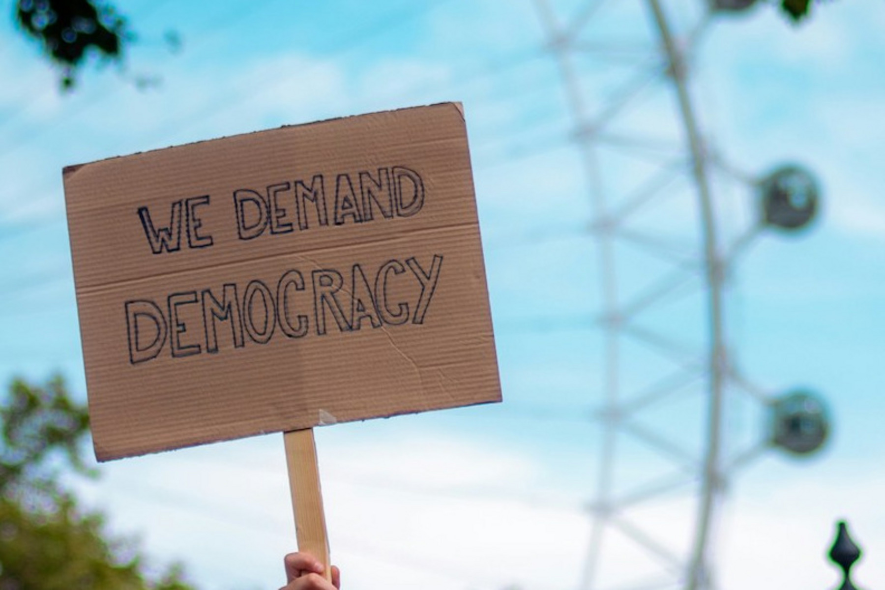 Das Bild zeigt eine Hand, die ein Pappschild mit der Aufschrift „WE DEMAND DEMOCRACY“ hochhält.