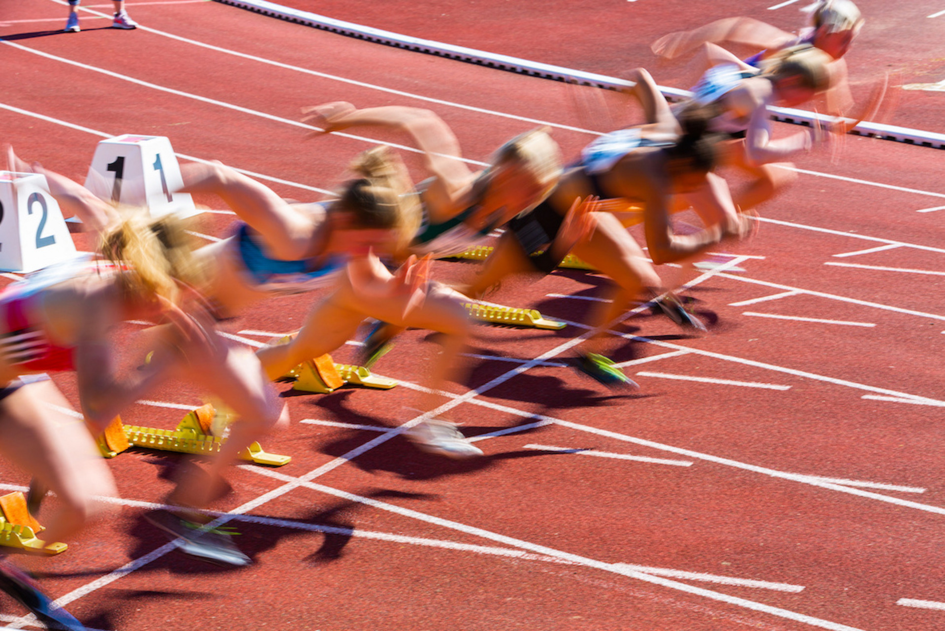 Startmoment eines Sprints bei einem Leichtathletik-Wettkampf. Mehrere Läuferinnen in Startposition auf einer roten Laufbahn, kurz nach dem Startschuss. Die Bewegung ist durch Bewegungsunschärfe festgehalten, was die Dynamik und Geschwindigkeit des Starts betont. Im Vordergrund sind die Startblöcke mit Nummern zu sehen.