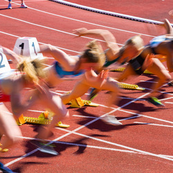 Startmoment eines Sprints bei einem Leichtathletik-Wettkampf. Mehrere Läuferinnen in Startposition auf einer roten Laufbahn, kurz nach dem Startschuss. Die Bewegung ist durch Bewegungsunschärfe festgehalten, was die Dynamik und Geschwindigkeit des Starts betont. Im Vordergrund sind die Startblöcke mit Nummern zu sehen.