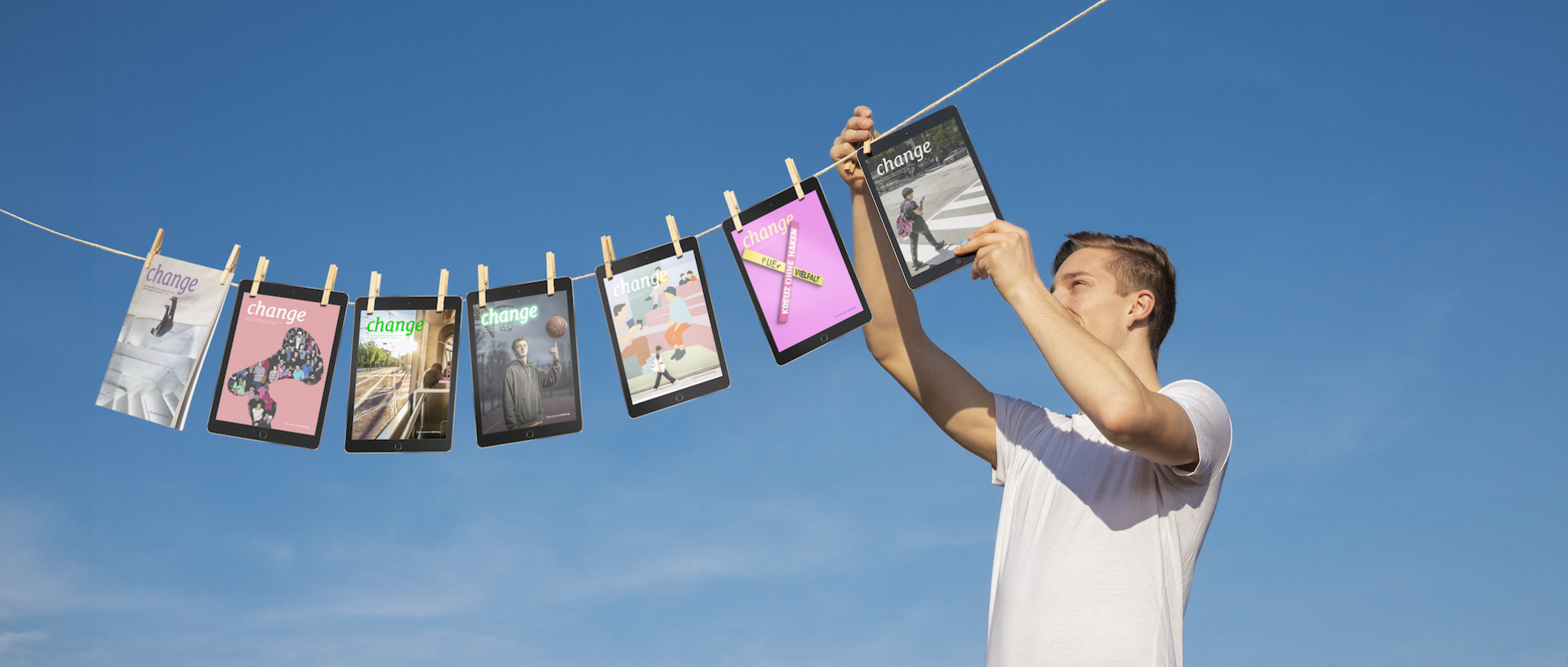 Vor blauem Himmel hängt eine Person in einem weißen T-Shirt verschiedene Ausgaben des 'change'-Magazins an eine Wäscheleine. Die Magazine sind mit Holzwäscheklammern befestigt und zeigen unterschiedliche Cover-Designs in verschiedenen Farben.