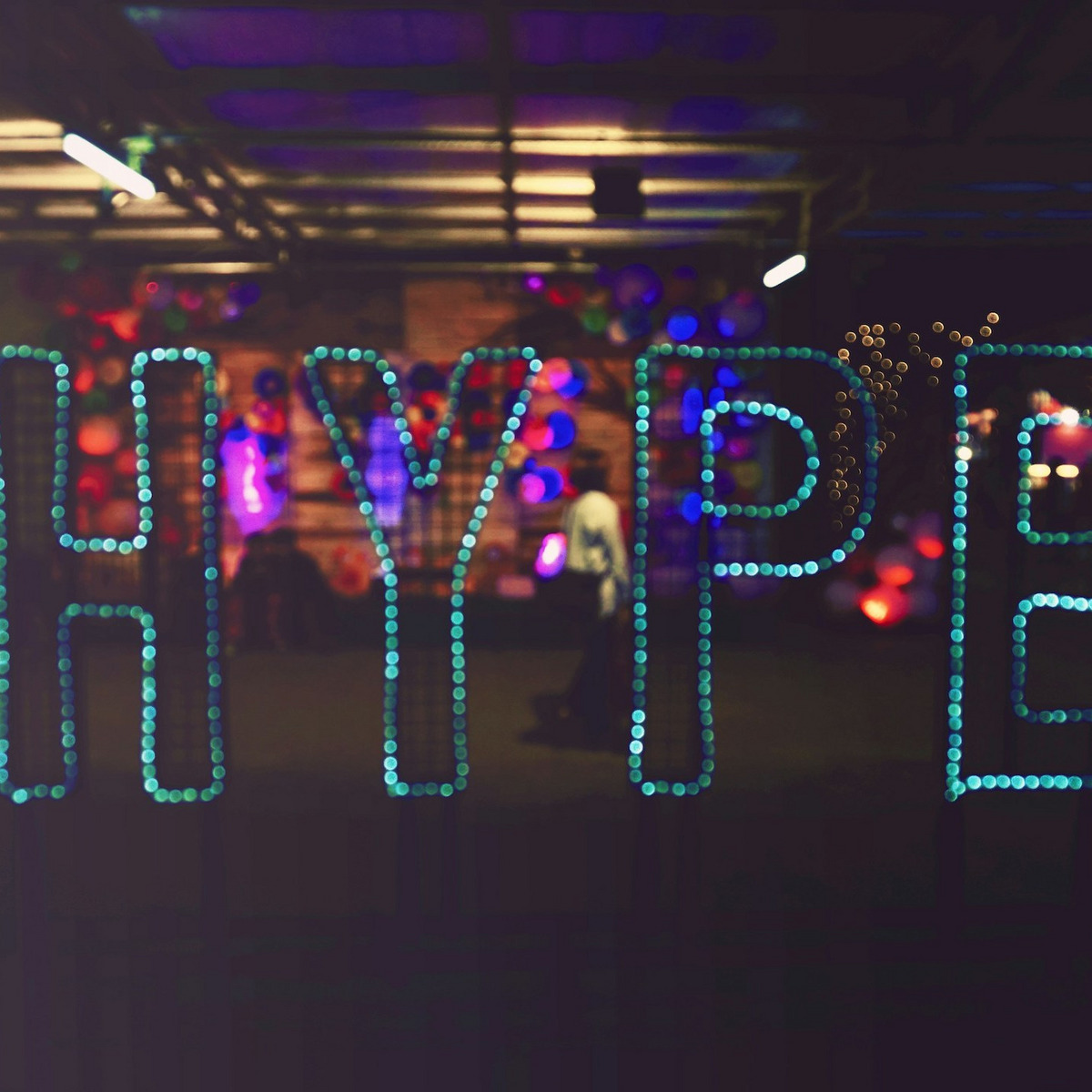 Das Wort 'HYPE' ist aus türkisfarbenen LED-Punkten oder kleinen Leuchten geformt und wird an einer Wand oder im Raum präsentiert. Im Hintergrund sind verschwommene bunte Lichter in Rot, Blau und Violett zu sehen.