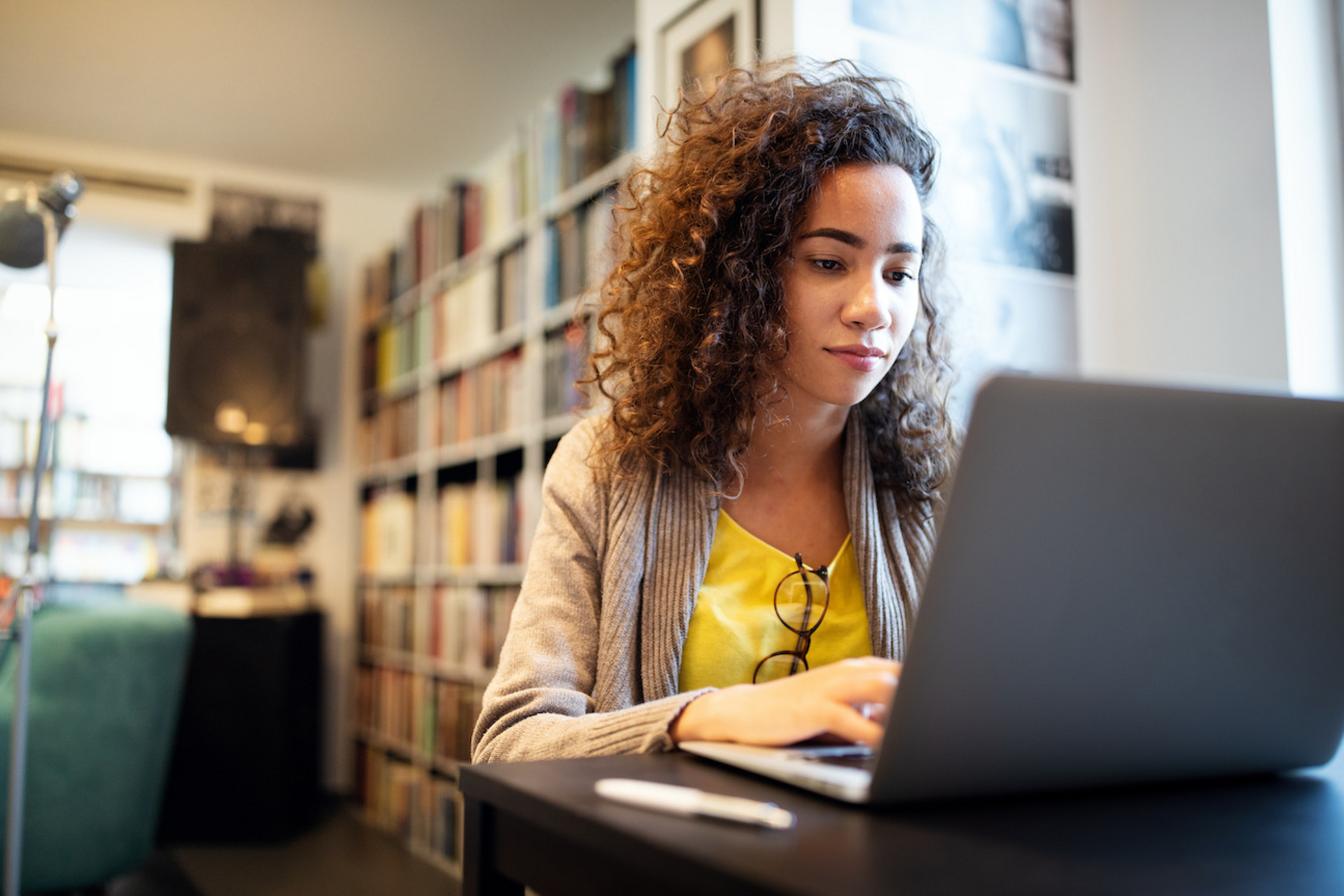 Junge Frau mit langen braunen Haaren sitzt lächelnd am Laptop. Sie trägt ein gelbes T-Shirt und eine graue Jacke. Im Hintergrund sind Bücherregale zu sehen.