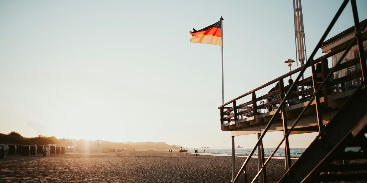 Das Bild zeigt einen Sandstrand bei Sonnenuntergang, auf dem im Vordergrund ein Gebäude auf Stelzen mit einer Treppe zu sehen ist. Eine deutsche Flagge weht auf einem hohen Fahnenmast neben dem Gebäude. Im Hintergrund sind Strandkörbe entlang des Strandes aufgereiht, und einige Menschen spazieren entlang der Wasserkante. Die Sonne steht tief am Himmel und taucht die Szene in ein warmes, goldenes Licht. Das Bild vermittelt eine ruhige und entspannte Atmosphäre.