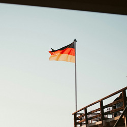 Das Bild zeigt einen Sandstrand bei Sonnenuntergang, auf dem im Vordergrund ein Gebäude auf Stelzen mit einer Treppe zu sehen ist. Eine deutsche Flagge weht auf einem hohen Fahnenmast neben dem Gebäude. Im Hintergrund sind Strandkörbe entlang des Strandes aufgereiht, und einige Menschen spazieren entlang der Wasserkante. Die Sonne steht tief am Himmel und taucht die Szene in ein warmes, goldenes Licht. Das Bild vermittelt eine ruhige und entspannte Atmosphäre.