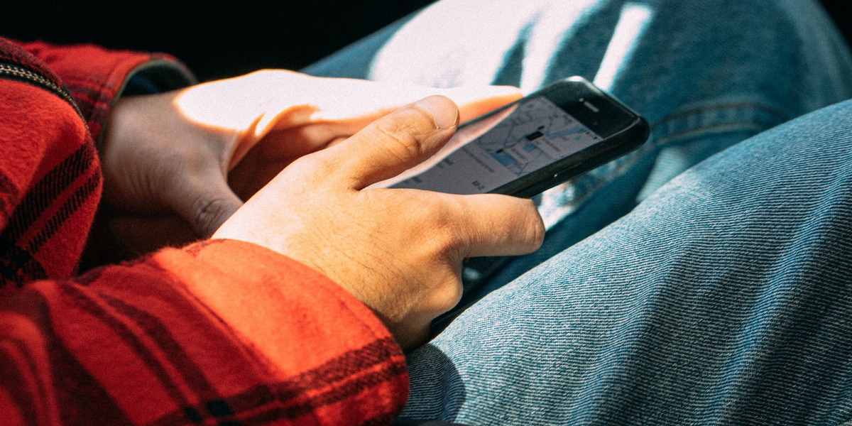 Warmes Sonnenlicht fällt durch die Scheibe eines Autos auf den Passagier. Wir sehen lediglich seine Hände, die ein Smartphone halten. Der Daumen ist zum Klick gehoben, auf dem Display ist eine Karte abgebildet.