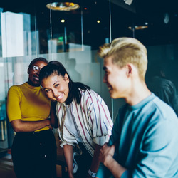 Drei junge Menschen stehen in einem modernen Büro und lachen zusammen. Die Person links trägt ein gelbes Oberteil und eine Brille, die Person in der Mitte hat dunkles Haar, das zu einem Pferdeschwanz gebunden ist, und trägt eine gestreifte Bluse. Die Person rechts hat kurzes blondes Haar und trägt ein hellblaues Oberteil. Im Hintergrund sind Glaswände und eine reflektierende Oberfläche zu sehen. Auf einem Tisch steht ein Kaffeebecher. Die Szene wirkt lebendig und fröhlich.