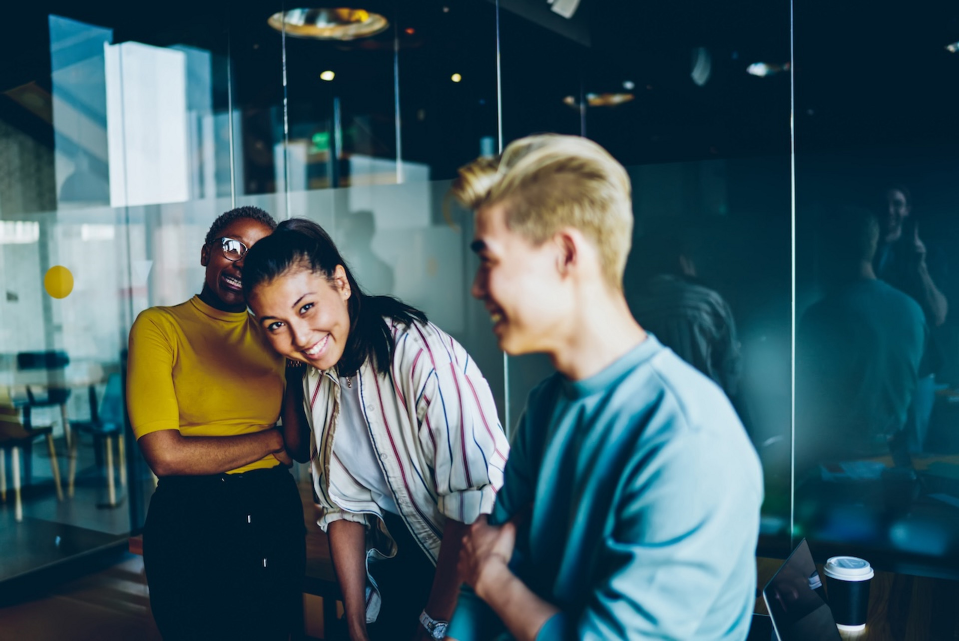 Drei junge Menschen stehen in einem modernen Büro und lachen zusammen. Die Person links trägt ein gelbes Oberteil und eine Brille, die Person in der Mitte hat dunkles Haar, das zu einem Pferdeschwanz gebunden ist, und trägt eine gestreifte Bluse. Die Person rechts hat kurzes blondes Haar und trägt ein hellblaues Oberteil. Im Hintergrund sind Glaswände und eine reflektierende Oberfläche zu sehen. Auf einem Tisch steht ein Kaffeebecher. Die Szene wirkt lebendig und fröhlich.