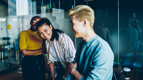 Drei junge Menschen stehen in einem modernen Büro und lachen zusammen. Die Person links trägt ein gelbes Oberteil und eine Brille, die Person in der Mitte hat dunkles Haar, das zu einem Pferdeschwanz gebunden ist, und trägt eine gestreifte Bluse. Die Person rechts hat kurzes blondes Haar und trägt ein hellblaues Oberteil. Im Hintergrund sind Glaswände und eine reflektierende Oberfläche zu sehen. Auf einem Tisch steht ein Kaffeebecher. Die Szene wirkt lebendig und fröhlich.