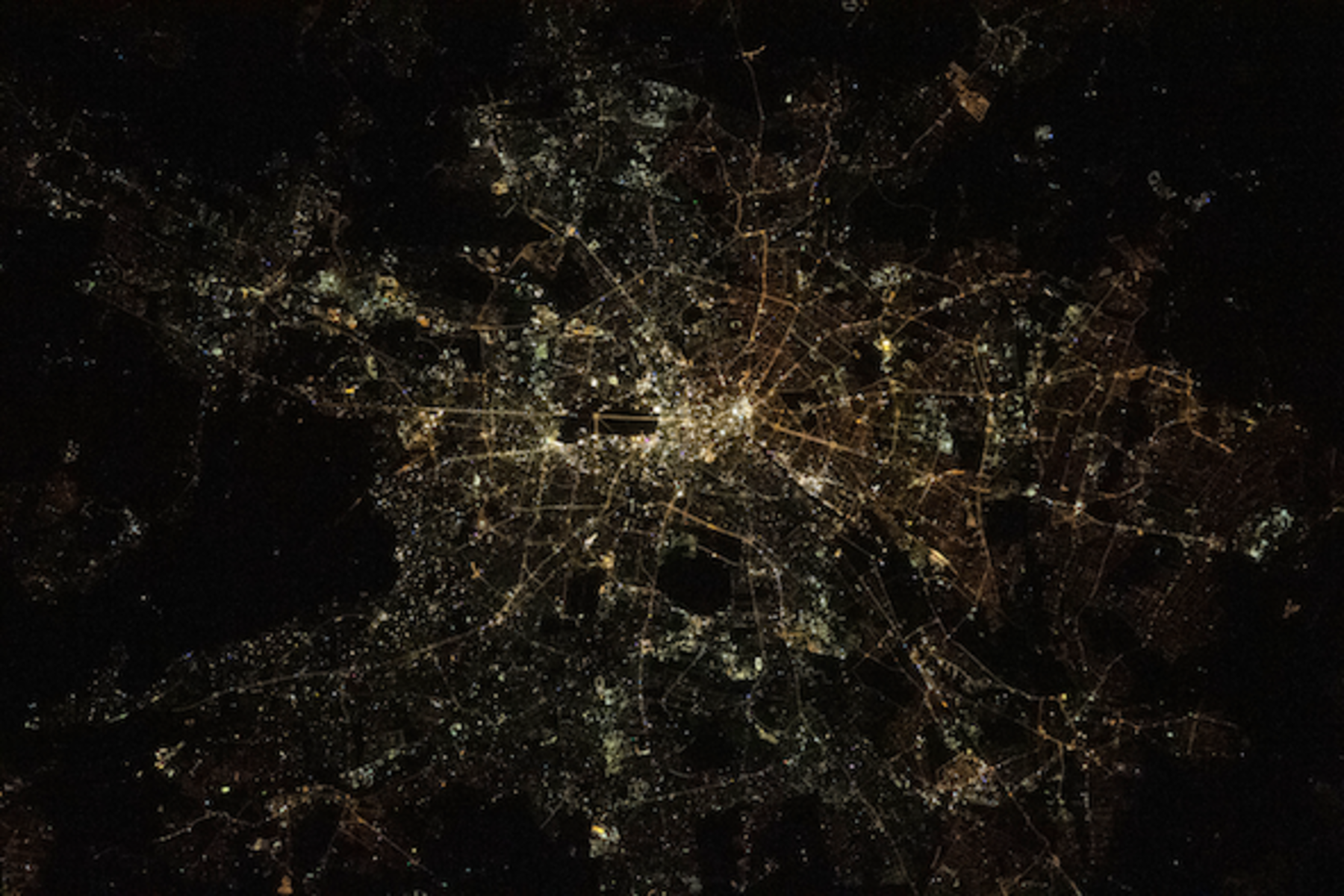 Das Bild zeigt eine nächtliche Luftaufnahme von Berlin, bei der die Stadt durch ihre Lichter sichtbar wird. Die hell erleuchteten Straßen und Gebäude bilden ein Netzwerk aus gelben und weißen Lichtpunkten, die das städtische Layout deutlich erkennen lassen. Der Stadtkern ist besonders hell, während sich die Lichter zu den Rändern der Stadt hin verdünnen. Dunklere Bereiche zwischen den Lichtern markieren Parks und unbebaute Flächen. Auffällig ist, dass die Lichter auf der linken Seite, also im Westen, eher weiß und gelb erscheinen, während sie auf der rechten Seite, im Osten, einen stärker orangefarbenen Ton aufweisen.