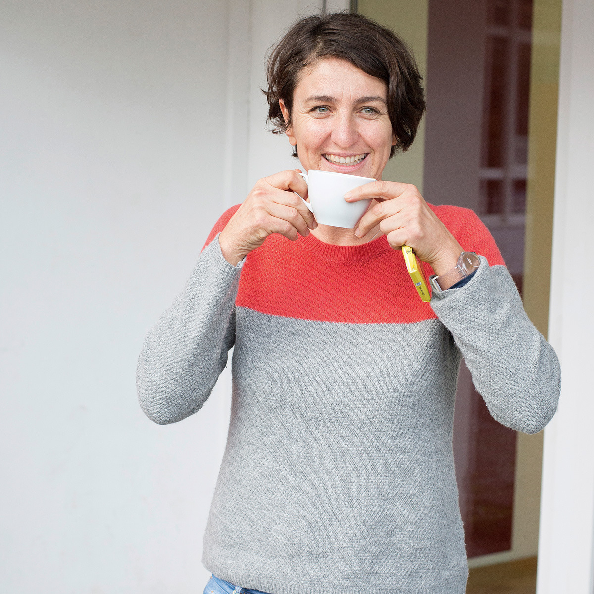 Christiane Falge trinkt aus einer Kaffeetasse und lacht in die Kamera.