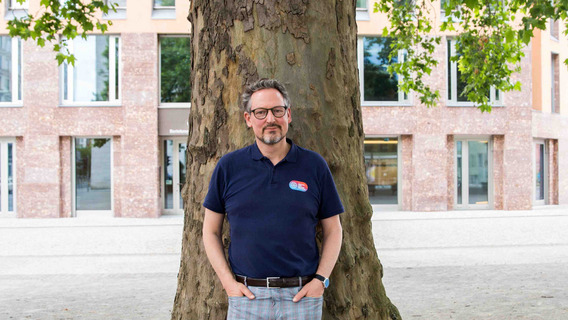 Eckart von Hirschhausen, ein Mann mittleren Alters, steht vor einem großen Baum. Er trägt eine Brille, ein dunkelblaues Polohemd mit einem kleinen Logo auf der linken Brust und eine karierte Hose. Seine Hände sind in den Hosentaschen. Im Hintergrund ist das Haus Berlin der Bertelsmann Stiftung, ein modernes Gebäude mit großen Fenstern und einer Fassade aus roten Ziegelsteinen, zu sehen. Der Mann wirkt entspannt und freundlich.