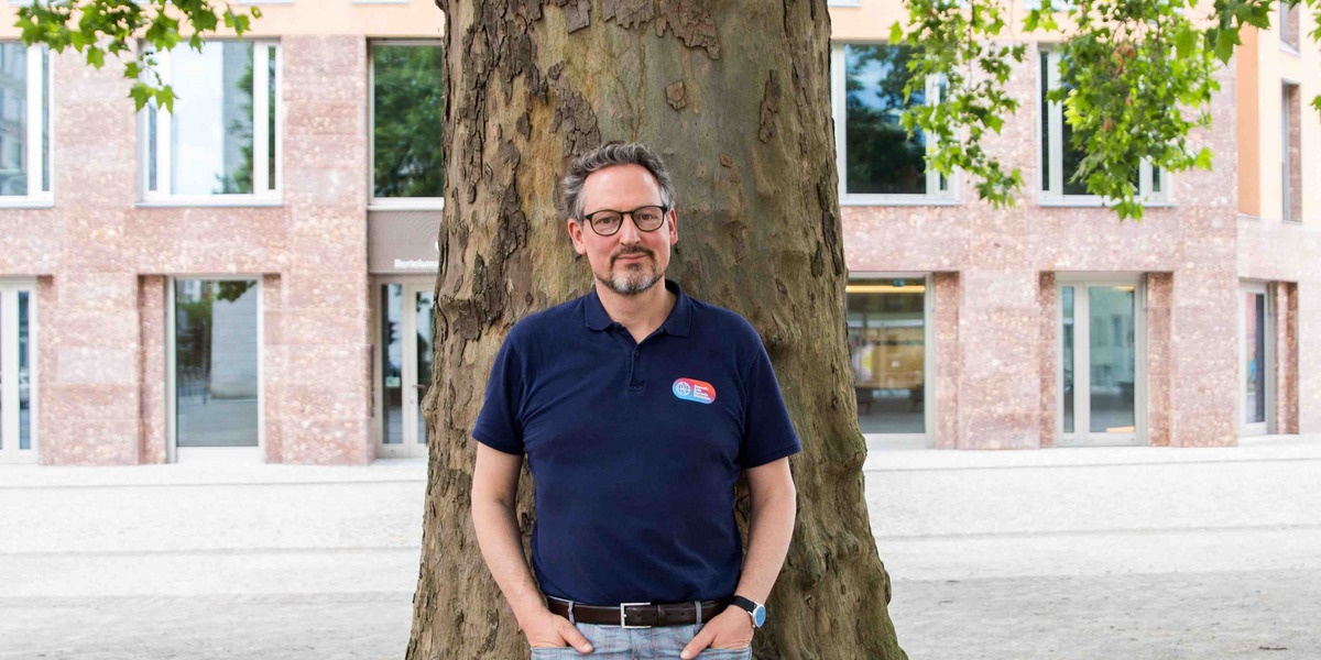 Eckart von Hirschhausen, ein Mann mittleren Alters, steht vor einem großen Baum. Er trägt eine Brille, ein dunkelblaues Polohemd mit einem kleinen Logo auf der linken Brust und eine karierte Hose. Seine Hände sind in den Hosentaschen. Im Hintergrund ist das Haus Berlin der Bertelsmann Stiftung, ein modernes Gebäude mit großen Fenstern und einer Fassade aus roten Ziegelsteinen, zu sehen. Der Mann wirkt entspannt und freundlich.