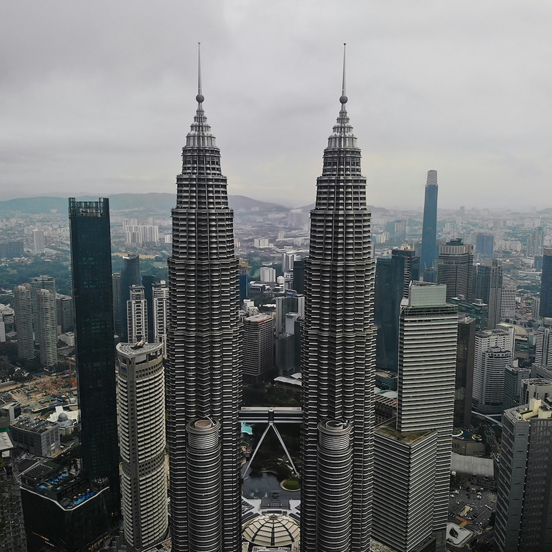 Im Vordergrund die Petronas Twin Towers, verbunden durch eine Brücke. Im Hintergrund ist die Skyline Kuala Lumpurs zu sehen, darüber ein wolkenverhangener, grauer Himmel.