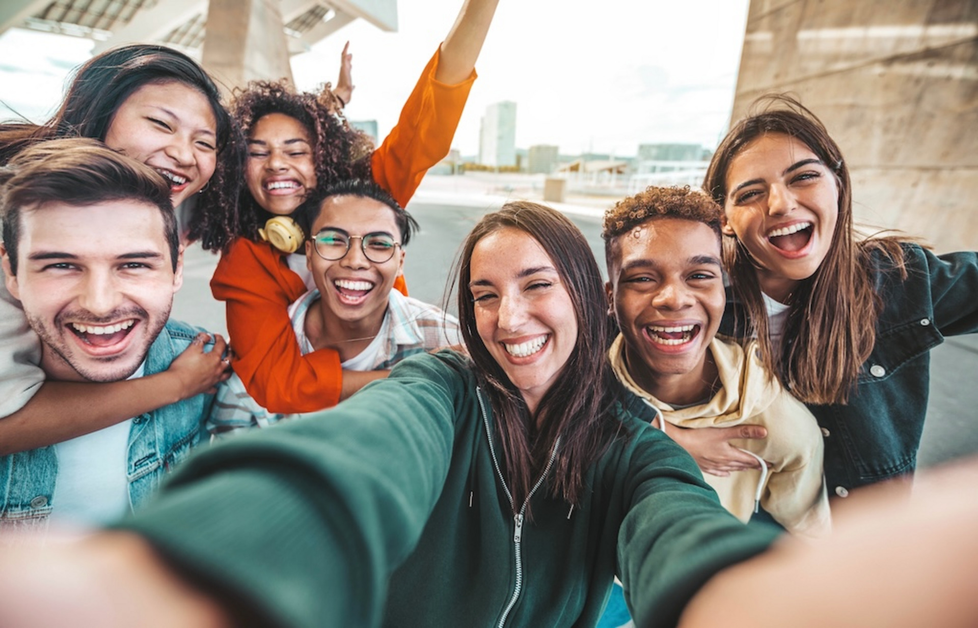 Eine Gruppe von sieben fröhlichen jungen Menschen macht ein Selfie, im Hintergrund ist die Kulisse einer Stadt zu sehen