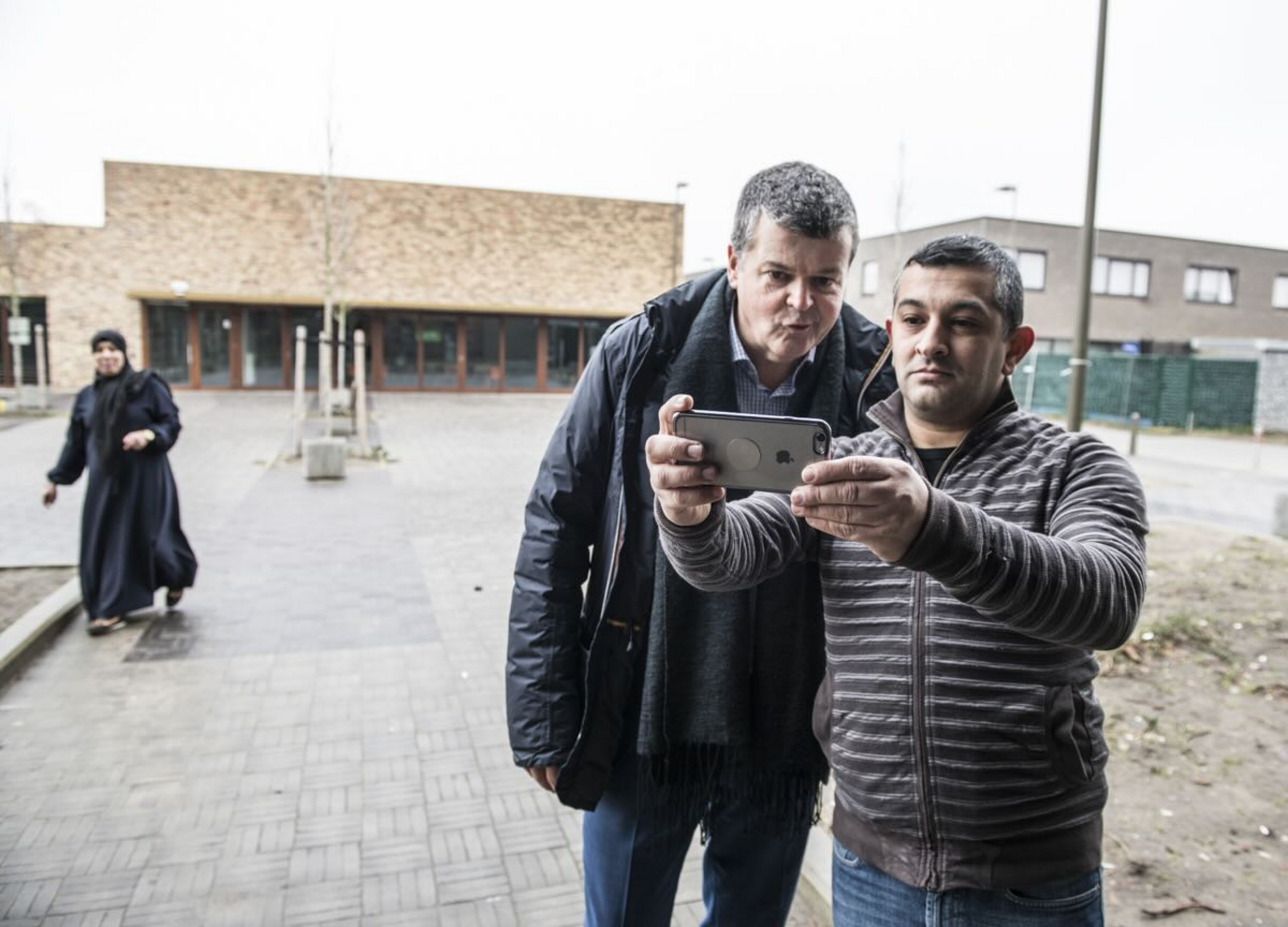 Zwei Männer stehen im Freien und machen ein Selfie mit einem Smartphone. Der Mann links ist der Bürgermeister von Mechelen, Bart Somers, er trägt eine dicke Jacke und einen Schal, während der rechte Mann eine gestreifte Jacke trägt. Im Hintergrund läuft eine Frau vorbei.