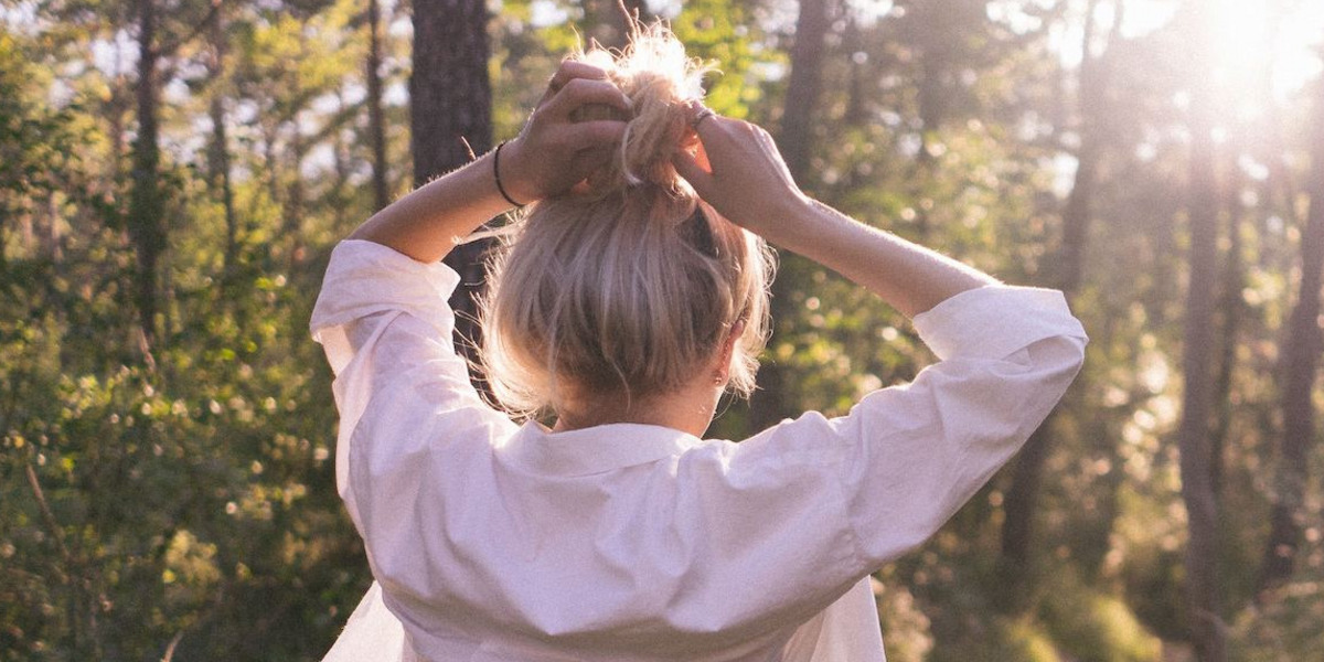 Eine junge Frau beim Spaziergang durch einen lichtdurchfluteten Wald, die Hände in die Haare greifend.