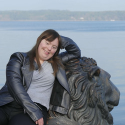 Luisa Wöllisch, eine Frau mit langen braunen Haaren und einem grauen Lederjackett, lehnt sich entspannt an eine Löwenstatue. Im Hintergrund sieht man einen ruhigen See und eine bewaldete Uferlinie. Luisa lächelt und wirkt glücklich.