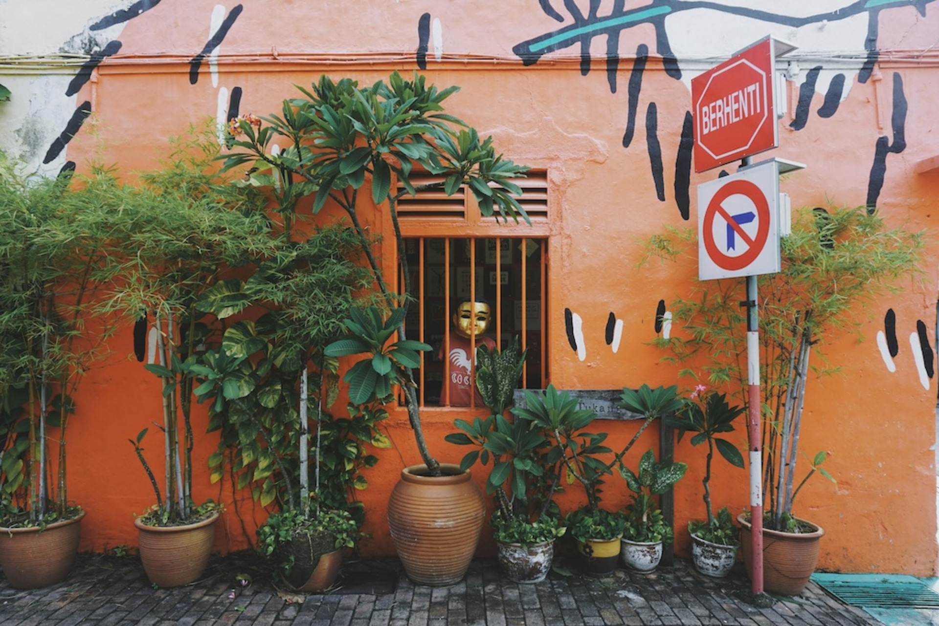 Das Bild zeigt eine farbenfrohe Szene in Melaka, Malaysia. Eine orangefarbene Wand mit künstlerischen schwarzen und weißen Akzenten bildet den Hintergrund. Im Vordergrund stehen mehrere grüne Pflanzen in Töpfen vor der Wand. Ein Fenster mit orangefarbenen Gittern befindet sich in der Mitte des Bildes, dahinter ist eine Figur mit einer goldenen Maske und einem roten Hemd zu sehen. Rechts neben dem Fenster sind zwei Straßenschilder zu erkennen: eines mit dem Wort „Berhenti“ (Malaiisch für „Stopp“), das andere zeigt ein Einfahrverbotsschild. Die gesamte Szene strahlt eine lebendige, urbane Atmosphäre aus.