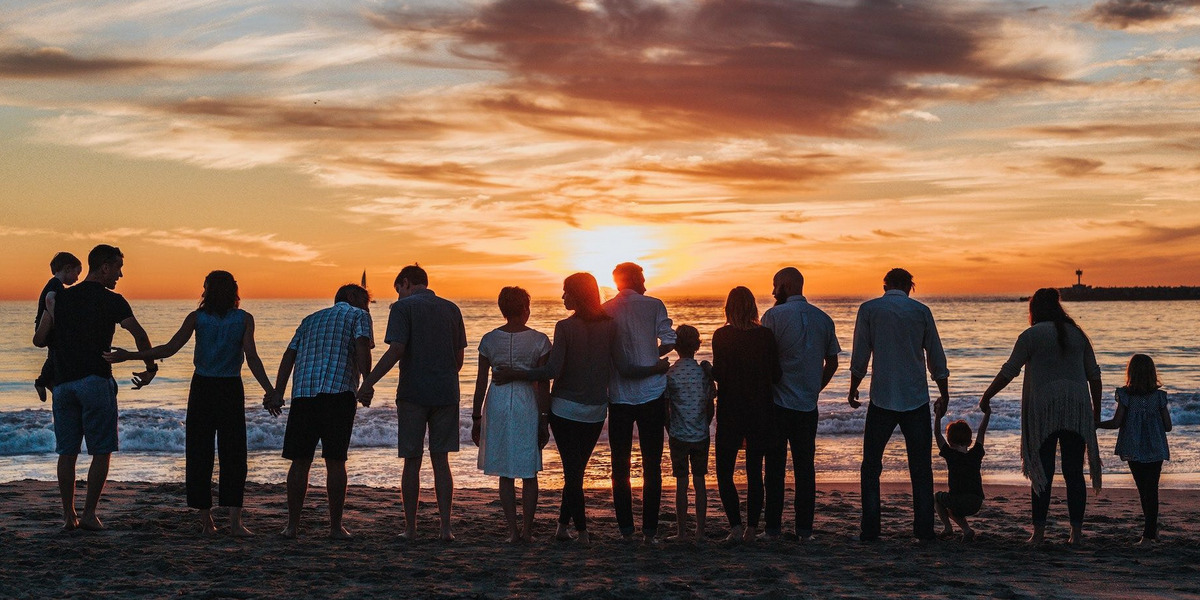 15 Menschen, aller Altersklassen und Geschlechter, stehen am Strand in einer Reihe und halten ihre Hände oder haben sich im Arm. Am Horizont geht die Sonne unter.