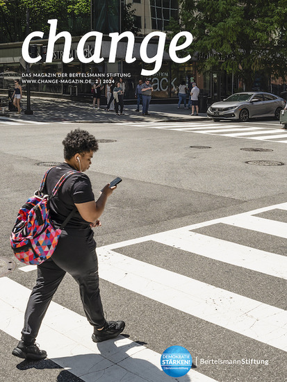 Titelseite des change Magazins der Bertelsmann Stiftung, Ausgabe 2/2024. Das Cover zeigt eine Person in schwarzer Kleidung mit einem bunten geometrisch gemusterten Rucksack, die auf ihr Smartphone schauend einen Zebrastreifen überquert.