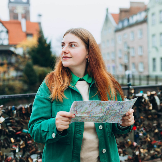 Eine rothaarige, junge Frau in grüner Jacke steht mit einem Stadtplan in der Hand auf einer Brücke in der polnischen Stadt Danzig und schaut sich um.