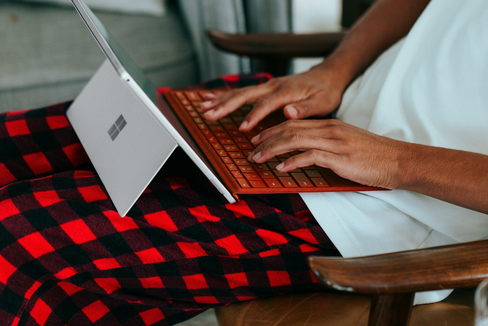 Nahaufnahme von Händen, die auf einer orangefarbenen Tastatur eines Microsoft Surface Laptops tippen. Der Laptop steht auf einer rot-schwarz karierten Decke, die über einem Holzstuhl liegt. Im Hintergrund ist der weiße Bildschirm des aufgeklappten Laptops zu sehen.