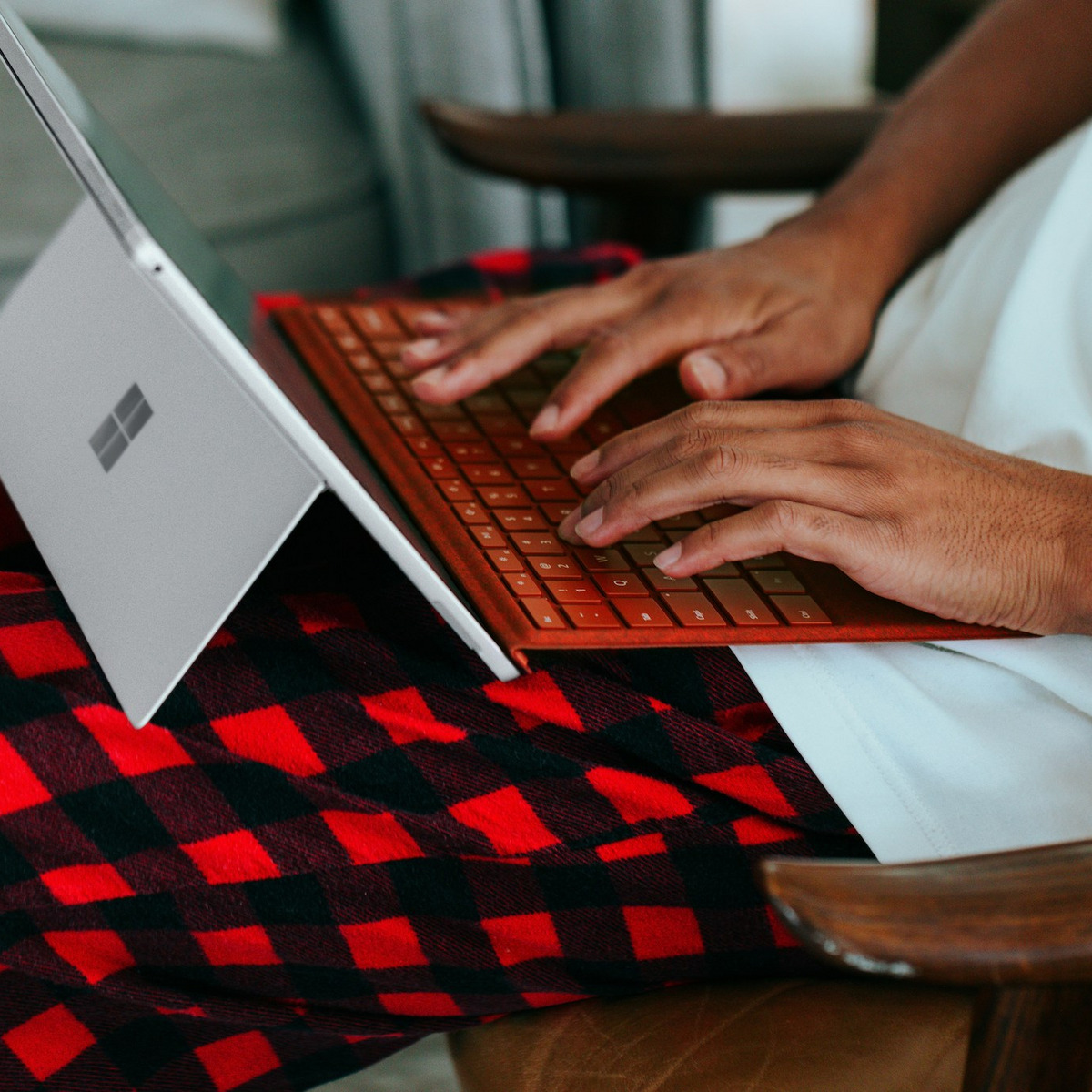 Nahaufnahme von Händen, die auf einer orangefarbenen Tastatur eines Microsoft Surface Laptops tippen. Der Laptop steht auf einer rot-schwarz karierten Decke, die über einem Holzstuhl liegt. Im Hintergrund ist der weiße Bildschirm des aufgeklappten Laptops zu sehen.