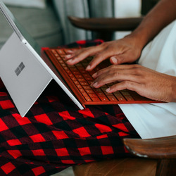 Nahaufnahme von Händen, die auf einer orangefarbenen Tastatur eines Microsoft Surface Laptops tippen. Der Laptop steht auf einer rot-schwarz karierten Decke, die über einem Holzstuhl liegt. Im Hintergrund ist der weiße Bildschirm des aufgeklappten Laptops zu sehen.