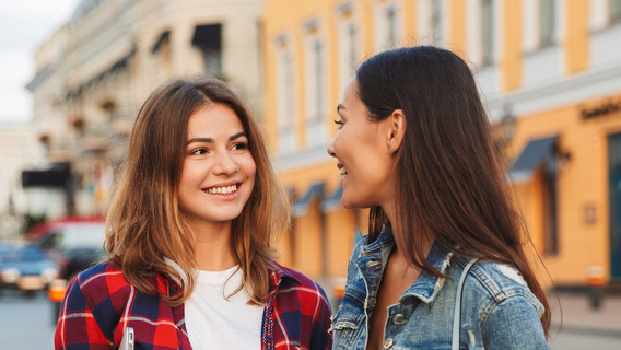 Zwei junge Frauen stehen auf einem Platz und lächeln sich an.