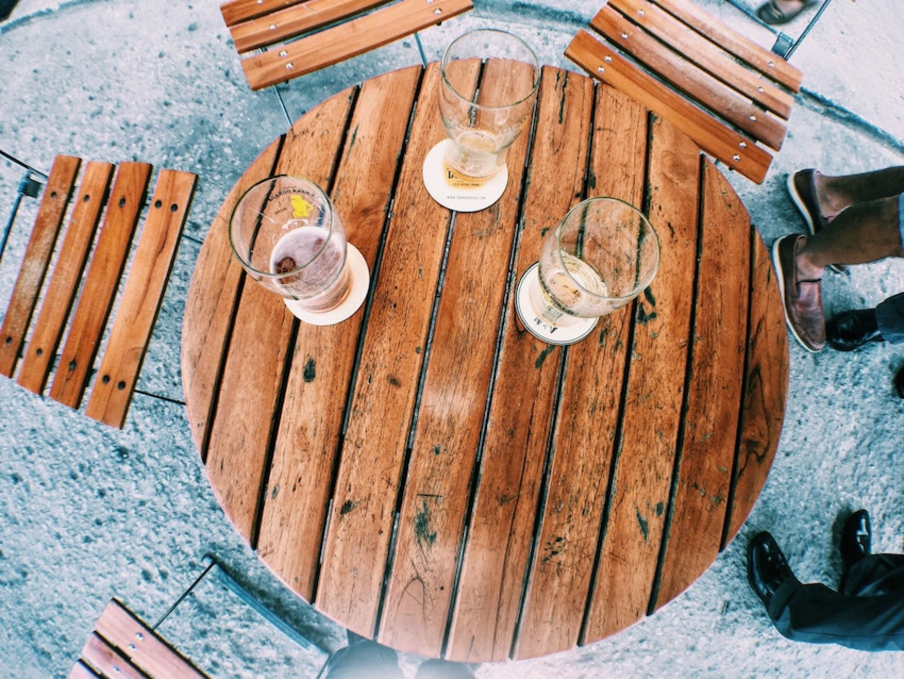 Ein runder Holztisch im Freien mit drei teils fast leeren, teils leeren Gläsern Bier auf Untersetzern, umgeben von klappbaren Holzstühlen. Es wirkt, als ob sich Personen um den Tisch herum befinden, erkennbar an den Füßen und Schuhen am Bildrand.