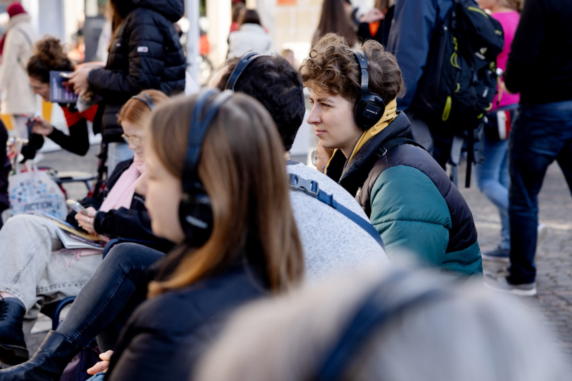 Eine belebte Außenszene mit sitzenden und stehenden Menschen. Im Vordergrund sitzen mehrere junge Leute mit Kopfhörern. Eine Person mit lockigem Haar und grün-schwarzer Jacke schaut konzentriert und trägt gelbe Kopfhörer. Andere um sie herum tragen ebenfalls Kopfhörer, was auf eine Audio-Tour oder -Veranstaltung hindeutet. Im Hintergrund sieht man einen geschäftigen Bereich mit weiteren gehenden und stehenden Menschen.