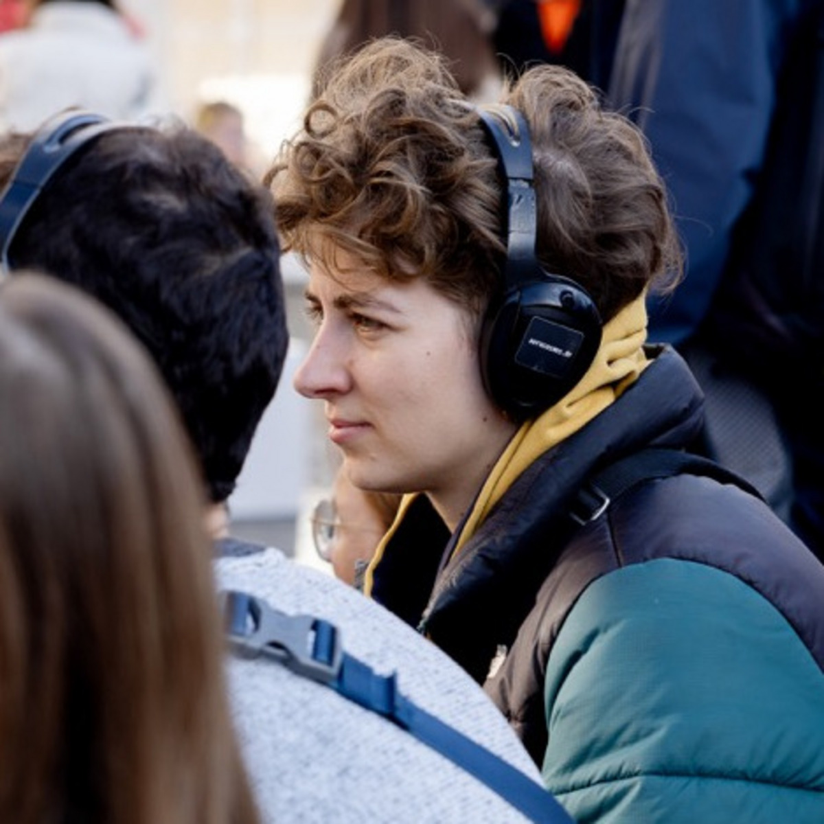 Eine belebte Außenszene mit sitzenden und stehenden Menschen. Im Vordergrund sitzen mehrere junge Leute mit Kopfhörern. Eine Person mit lockigem Haar und grün-schwarzer Jacke schaut konzentriert und trägt gelbe Kopfhörer. Andere um sie herum tragen ebenfalls Kopfhörer, was auf eine Audio-Tour oder -Veranstaltung hindeutet. Im Hintergrund sieht man einen geschäftigen Bereich mit weiteren gehenden und stehenden Menschen.