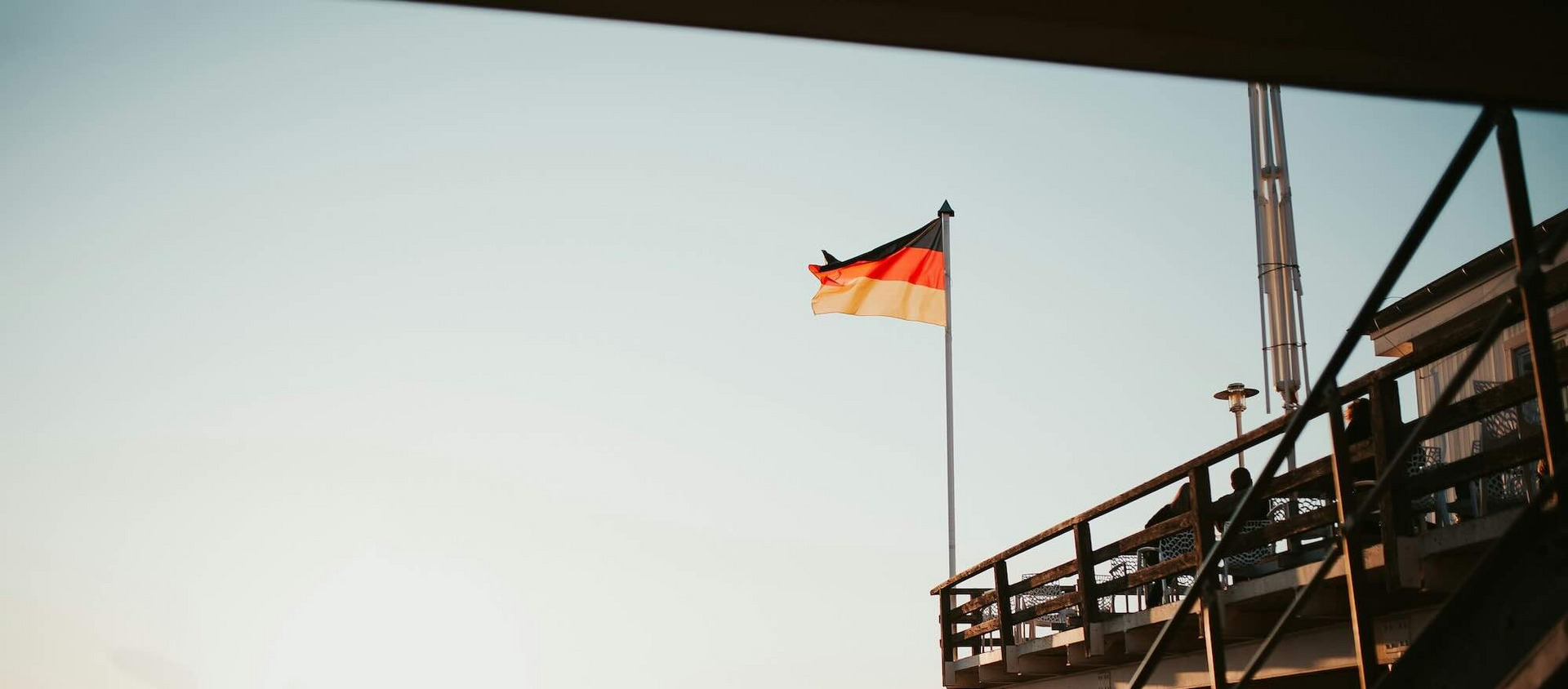 Das Bild zeigt einen Sandstrand bei Sonnenuntergang, auf dem im Vordergrund ein Gebäude auf Stelzen mit einer Treppe zu sehen ist. Eine deutsche Flagge weht auf einem hohen Fahnenmast neben dem Gebäude. Im Hintergrund sind Strandkörbe entlang des Strandes aufgereiht, und einige Menschen spazieren entlang der Wasserkante. Die Sonne steht tief am Himmel und taucht die Szene in ein warmes, goldenes Licht. Das Bild vermittelt eine ruhige und entspannte Atmosphäre.