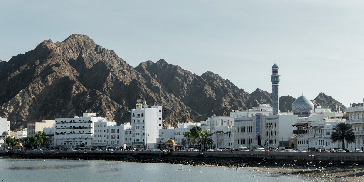 Das Bild zeigt eine Küstenstadt, die von felsigen Bergen umgeben ist. Die Stadt ist eine Mischung aus traditioneller arabischer Architektur, einschließlich einer prominenten Moschee mit Minarett, sowie modernen Hochhäusern. Die Küstenlinie ist mit Palmen gesäumt und es sind kleine Boote im Wasser zu sehen. Die Gesamtszene vermittelt eine malerische, am Meer gelegene Umgebung mit einer Mischung aus alten und neuen Elementen. Ohne dass konkrete Personen identifiziert werden können, deutet das Bild auf eine lebendige, kulturell reiche Küstenstadt in einer Region des Nahen Ostens oder Nordafrikas hin.