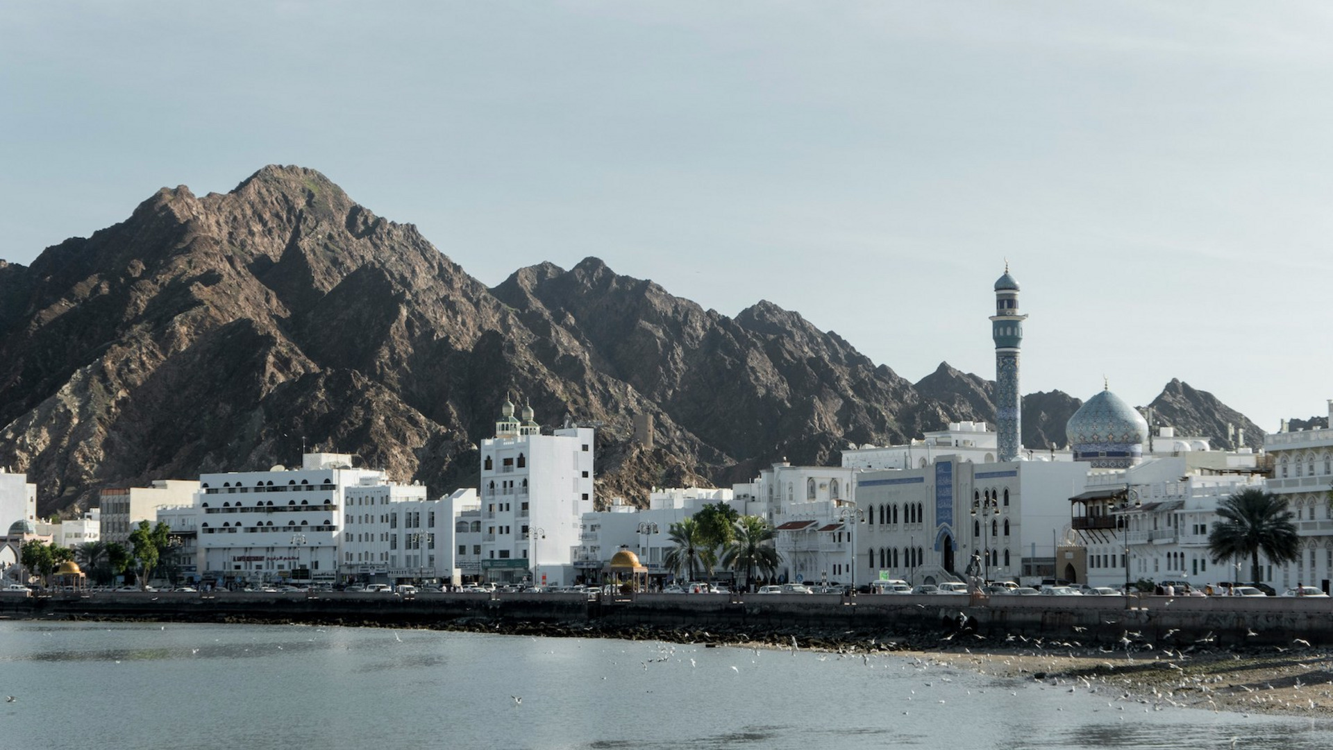 Das Bild zeigt eine Küstenstadt, die von felsigen Bergen umgeben ist. Die Stadt ist eine Mischung aus traditioneller arabischer Architektur, einschließlich einer prominenten Moschee mit Minarett, sowie modernen Hochhäusern. Die Küstenlinie ist mit Palmen gesäumt und es sind kleine Boote im Wasser zu sehen. Die Gesamtszene vermittelt eine malerische, am Meer gelegene Umgebung mit einer Mischung aus alten und neuen Elementen. Ohne dass konkrete Personen identifiziert werden können, deutet das Bild auf eine lebendige, kulturell reiche Küstenstadt in einer Region des Nahen Ostens oder Nordafrikas hin.