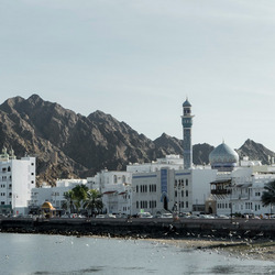 Das Bild zeigt eine Küstenstadt, die von felsigen Bergen umgeben ist. Die Stadt ist eine Mischung aus traditioneller arabischer Architektur, einschließlich einer prominenten Moschee mit Minarett, sowie modernen Hochhäusern. Die Küstenlinie ist mit Palmen gesäumt und es sind kleine Boote im Wasser zu sehen. Die Gesamtszene vermittelt eine malerische, am Meer gelegene Umgebung mit einer Mischung aus alten und neuen Elementen. Ohne dass konkrete Personen identifiziert werden können, deutet das Bild auf eine lebendige, kulturell reiche Küstenstadt in einer Region des Nahen Ostens oder Nordafrikas hin.