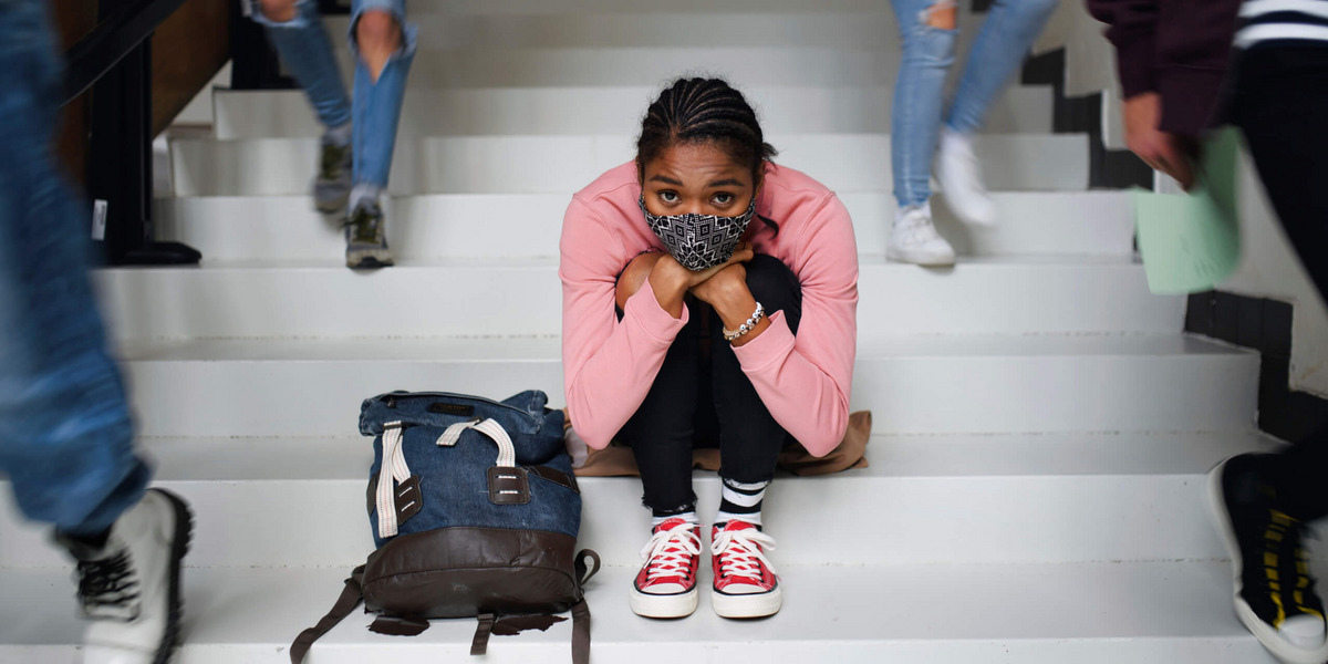 Eine junge Frau mit Alltagsmaske sitzt auf den Treppenstufen in der Schule und schaut besorgt. Neben ihr laufen Schulkamerad:innen die Treppe hinab.