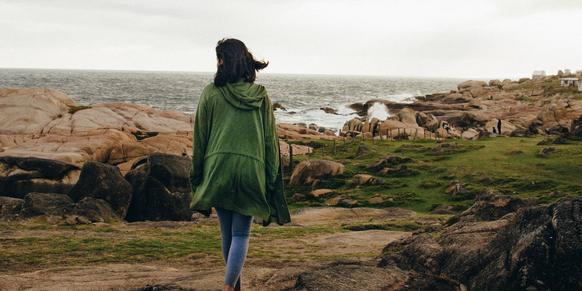 Eine Person in einem grünen Mantel läuft, uns den Rücken zugekehrt, den felsigen Strand von Cabo Polonio in Uruguay entlang, auf den tosende Wellen treffen.