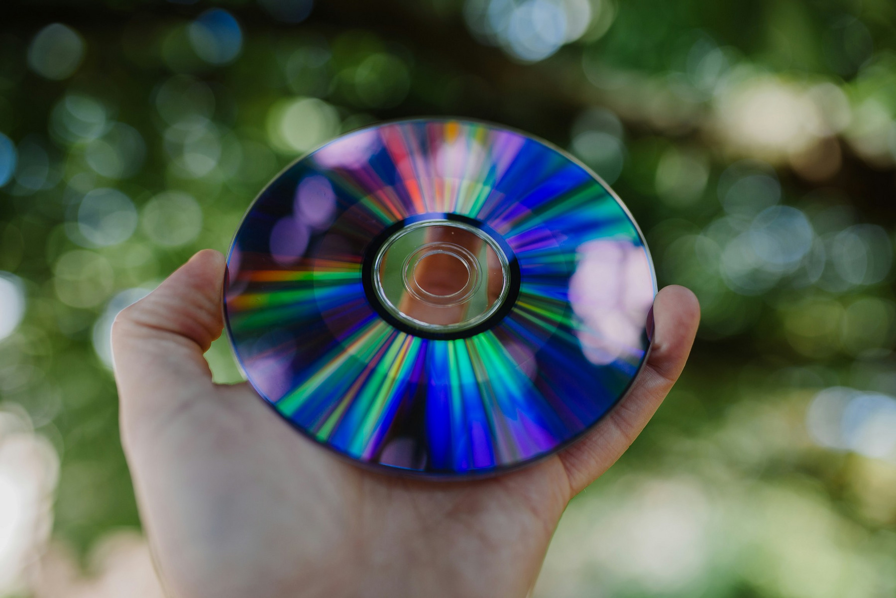 Eine Hand hält eine DVD, die das Sonnenlicht einfängt und bunte Regenbogenfarben reflektiert. Der Hintergrund ist unscharf und zeigt grüne, verschwommene Blätter, was auf eine Aufnahme im Freien hindeutet.