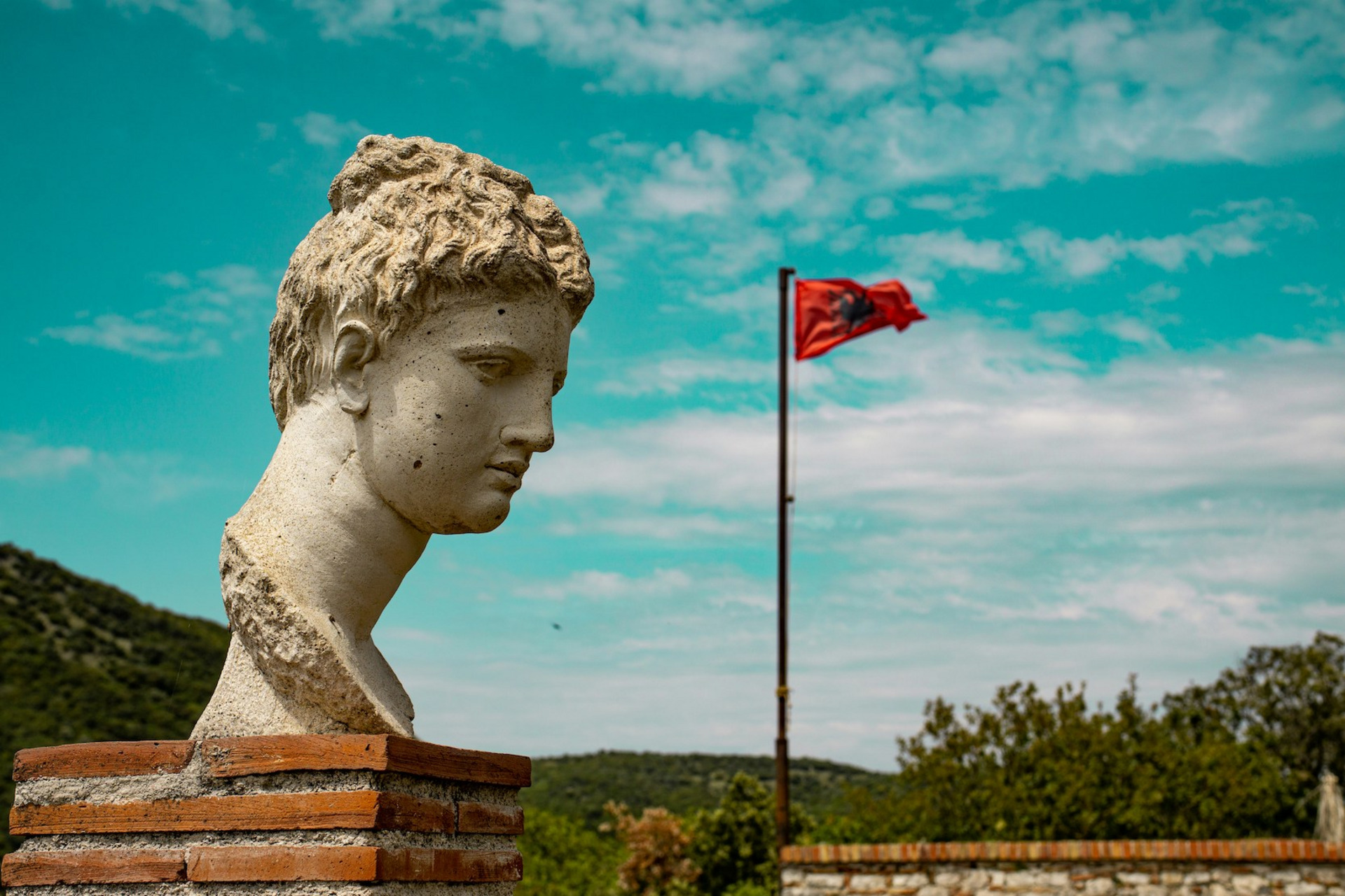 : Zu sehen sind der Kopf einer antiken Statue vor einer mediterranen Hügellandschaft im Nationalpark Butrint. Im Hintergrund weht die albanische Flagge.