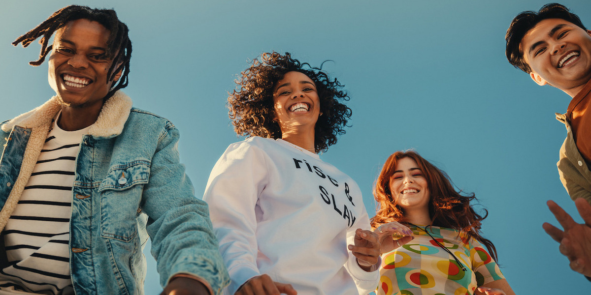 Eine Gruppe von vier fröhlichen, lachenden jungen Menschen, die vor einem blauen Himmelshintergrund stehen. Sie tragen bunte, lässige Kleidung wie Jeans, Pullover und T-Shirts. Ihre Körpersprache und Gesichtsausdrücke signalisieren eine ausgelassene, lebendige Stimmung