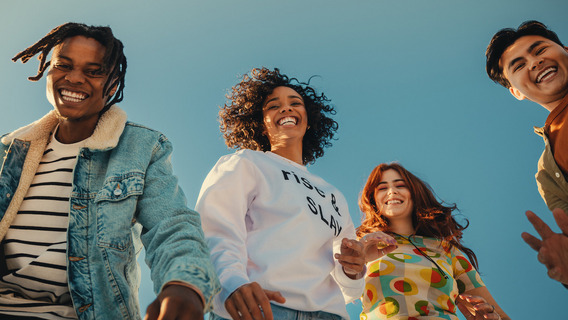 Eine Gruppe von vier fröhlichen, lachenden jungen Menschen, die vor einem blauen Himmelshintergrund stehen. Sie tragen bunte, lässige Kleidung wie Jeans, Pullover und T-Shirts. Ihre Körpersprache und Gesichtsausdrücke signalisieren eine ausgelassene, lebendige Stimmung