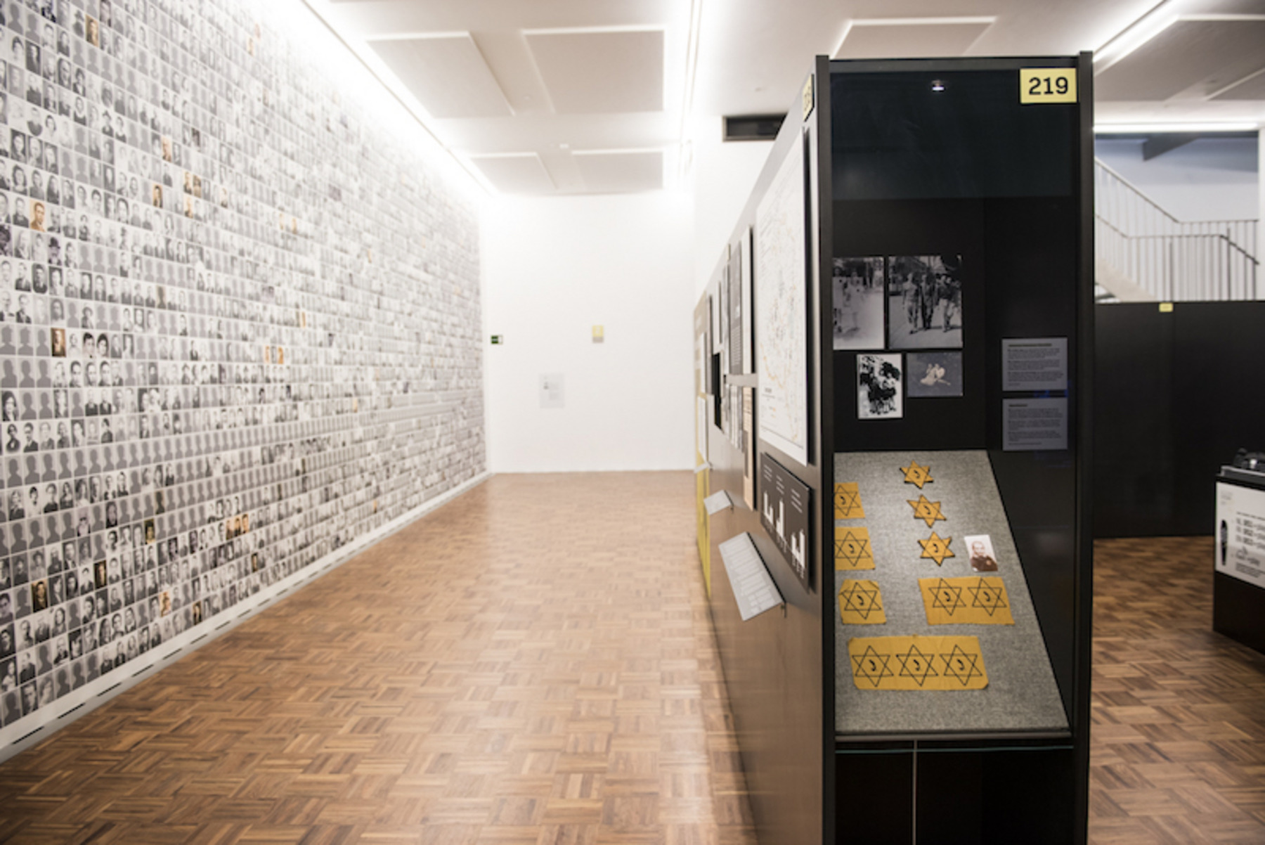 Ein Museumsausstellungsraum: Links ist eine lange Wand mit Porträts von Menschen zu sehen, und rechts in einer Vitrine liegen gelbe Davidsterne ausgestellt, wie sie während des Holocausts getragen wurden. Dieses Bild wurde in der Kaserne Dossin, dem Holocaust- und Menschenrechtsmuseum in Mechelen, aufgenommen.