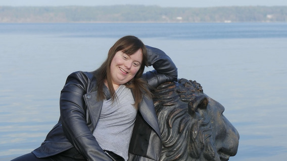 Luisa Wöllisch, eine Frau mit langen braunen Haaren und einem grauen Lederjackett, lehnt sich entspannt an eine Löwenstatue. Im Hintergrund sieht man einen ruhigen See und eine bewaldete Uferlinie. Luisa lächelt und wirkt glücklich.