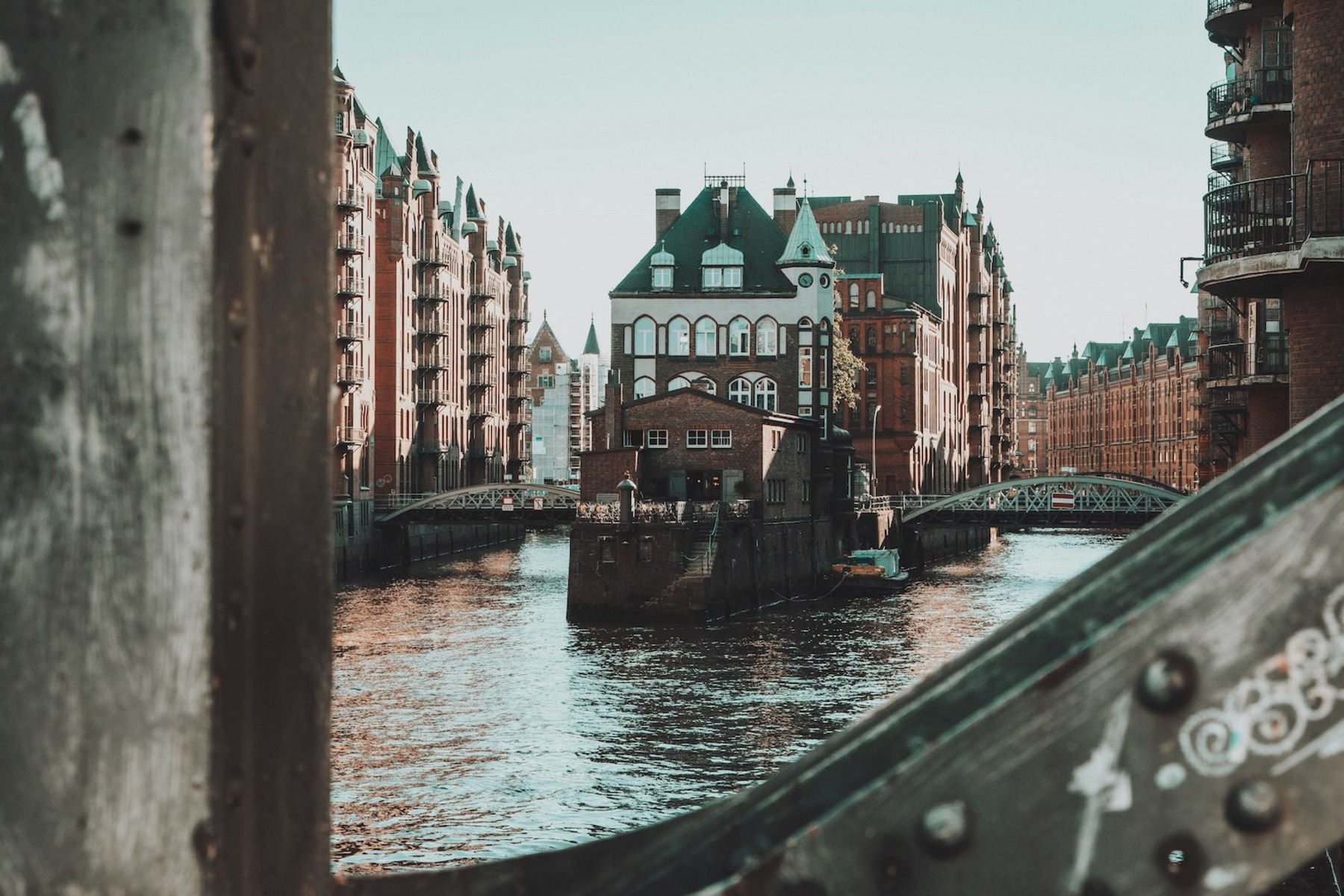 Das Bild zeigt die Hamburger Speicherstadt mit ihren charakteristischen roten Backsteingebäuden, die sich entlang eines Kanals erstrecken. Im Zentrum des Bildes steht ein historisches Gebäude, das von zwei Brücken flankiert wird. Das Bild wurde durch eine Öffnung in einer Brücke aufgenommen, wodurch der Rahmen des Bildes einen rustikalen, industriellen Touch erhält. Das Wasser im Kanal reflektiert das Licht des klaren Himmels.