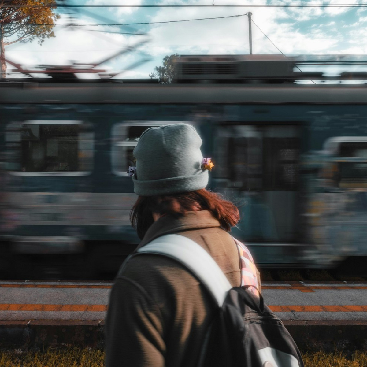 Eine Person mit grauer Mütze und Rucksack steht mit dem Rücken zur Kamera, im Hintergrund fährt ein Zug vorbei