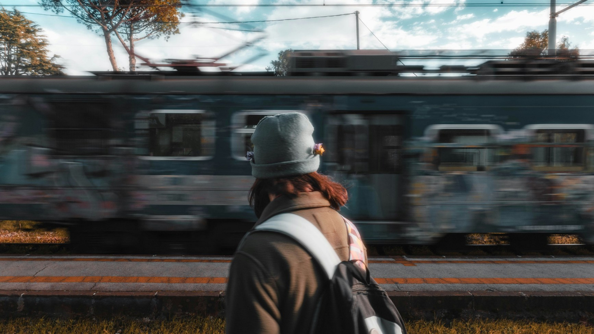 Eine Person mit grauer Mütze und Rucksack steht mit dem Rücken zur Kamera, im Hintergrund fährt ein Zug vorbei