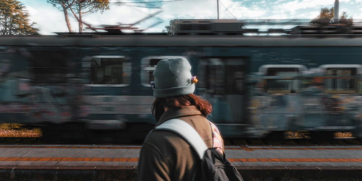 Eine Person mit grauer Mütze und Rucksack steht mit dem Rücken zur Kamera, im Hintergrund fährt ein Zug vorbei